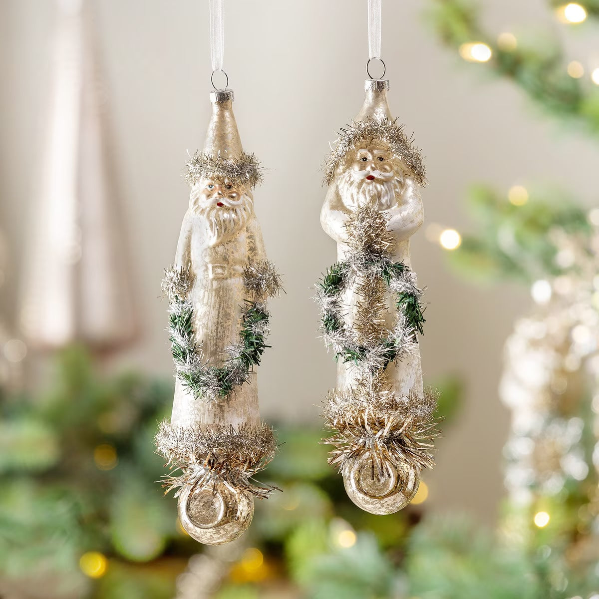 Two vintage-style Christmas ornaments shaped like Santa Claus hang from a tree. The ornaments are metallic silver with green tinsel garlands and white beards, set against a softly blurred holiday background with warm lights.